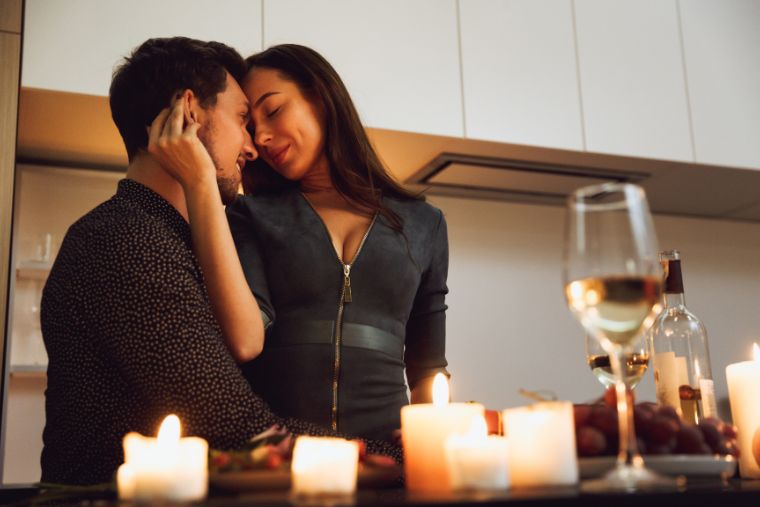 A woman with closed eyes rests her head on a man's forehead as she sits on his lap, near lit candles and wine.