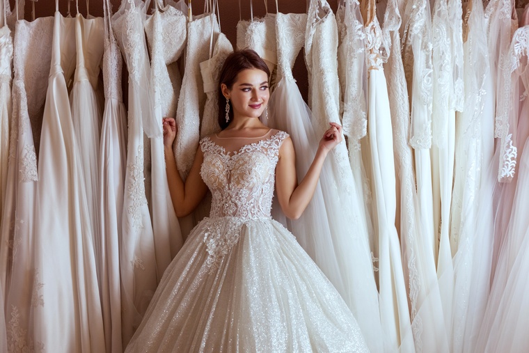 A woman in a white wedding dress leans back against dozens of other white gowns hanging on a rack in a boutique.