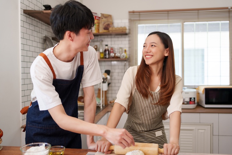 A happy pair of young people in kitchen aprons working together to make bread by rolling and flouring dough.