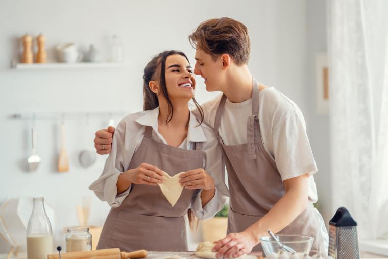 A smiling couple bakes together in a bright kitchen with baking tools and ingredients spread across the counter.