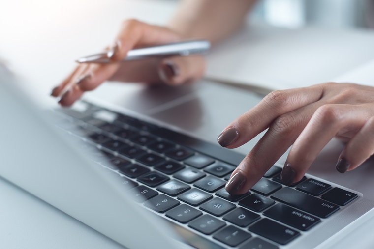 A person with painted fingernails typing on the used keyboard of a silver laptop while also holding a silver pen.