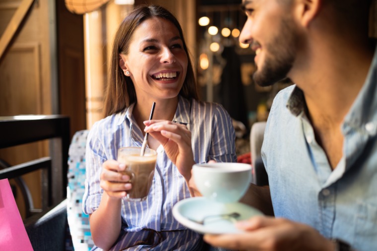 A smiling woman and a man sit close together at a café table near a large window. They hold their coffee beverages.