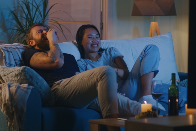 A diverse couple relaxing on a couch at night, surrounded by candles and wine, laughing together while watching a movie on TV.