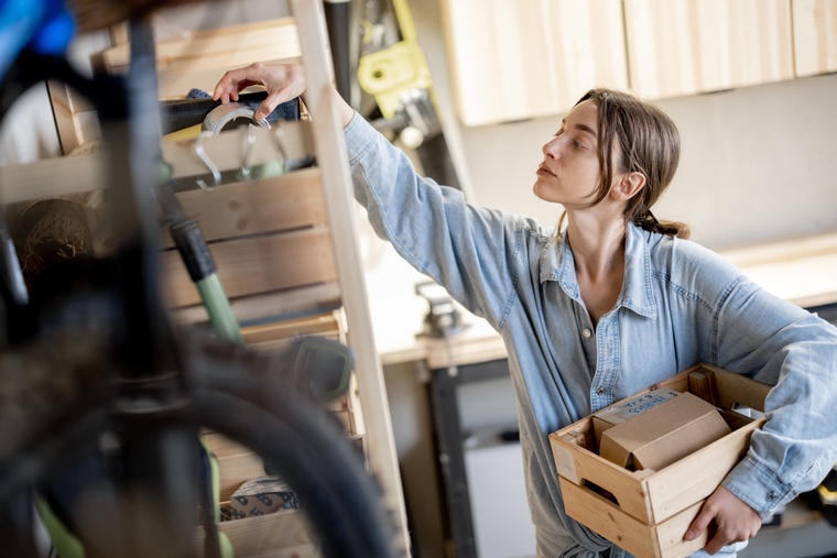 A young person pulling items off of a garage shelf and moving them into a small wooden bin tucked under her arm.