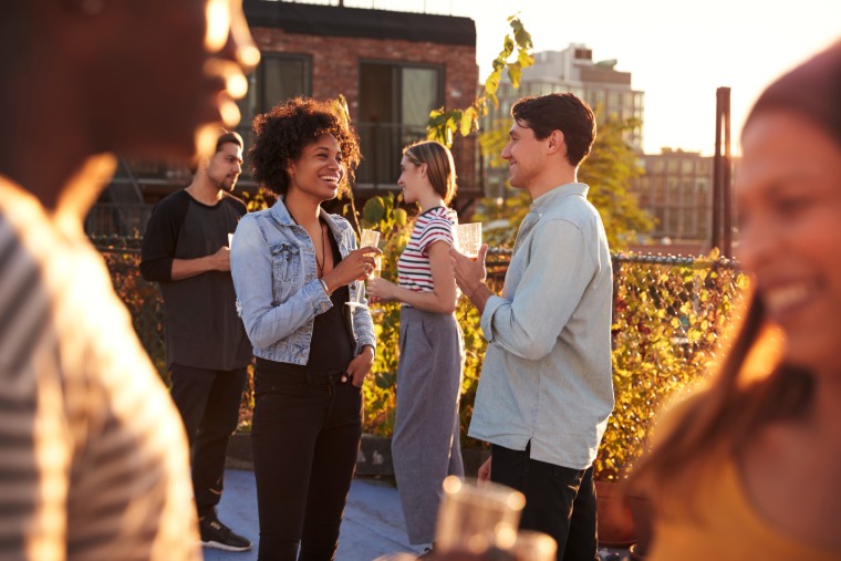 A diverse group of young adults chat at a rooftop singles mixer, holding drinks and smiling during golden hour.