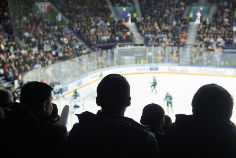 Hockey players on the ice in an indoor rink as seen over the shoulders of audience members watching the game.