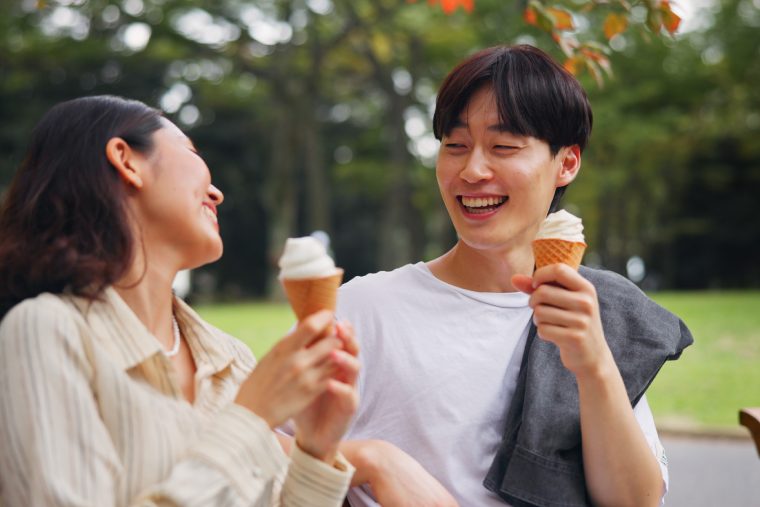 A young couple sitting on a park bench outside and enjoying vanilla ice cream cones together while smiling.