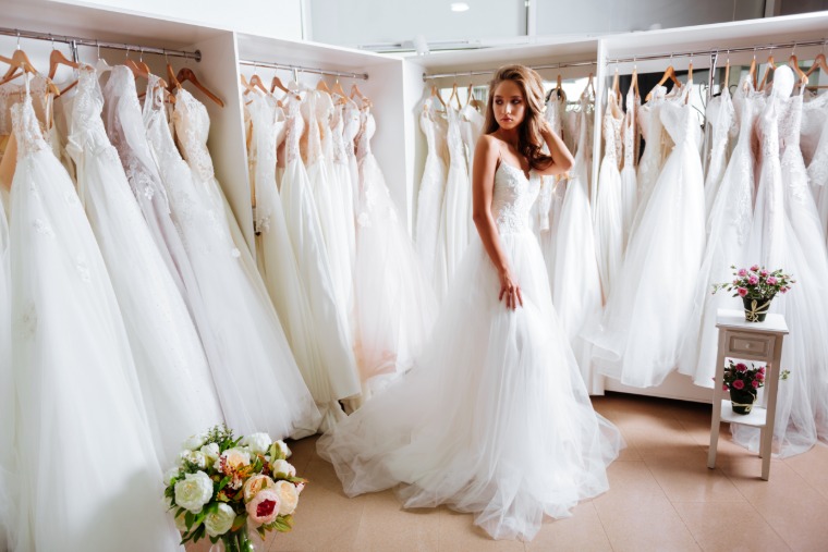 A woman in a wedding dress standing with her back turned while looking at bridal gowns inside a boutique.